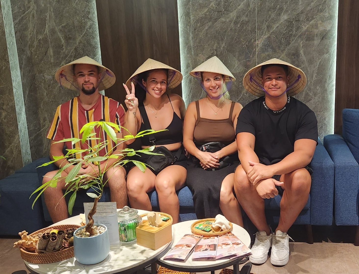 Two happy couples are smiling while wearing Vietnamese conical hats and sitting on a blue sofa in a spa.