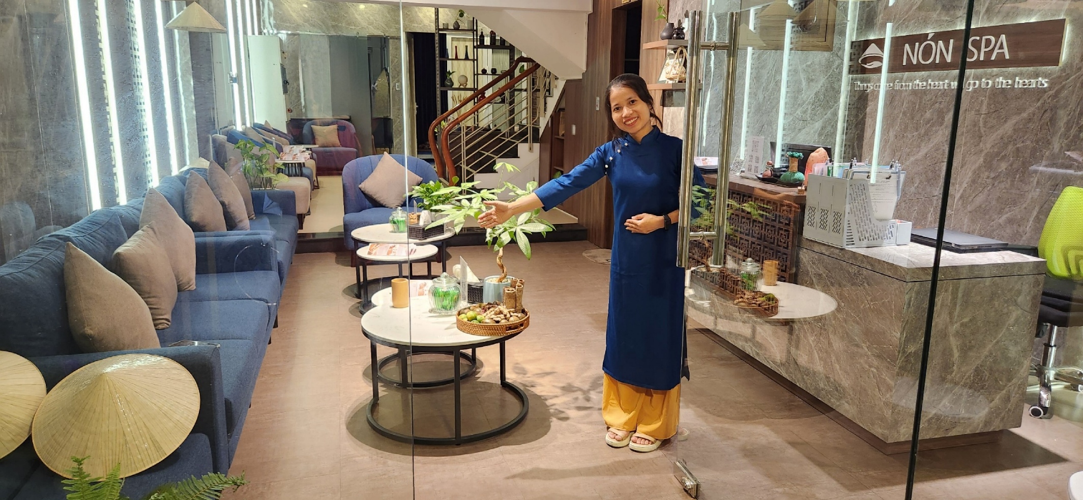 A spa receptionist wearing Vietnamese's "Ao dai" is welcoming customers next to an opening glass door.