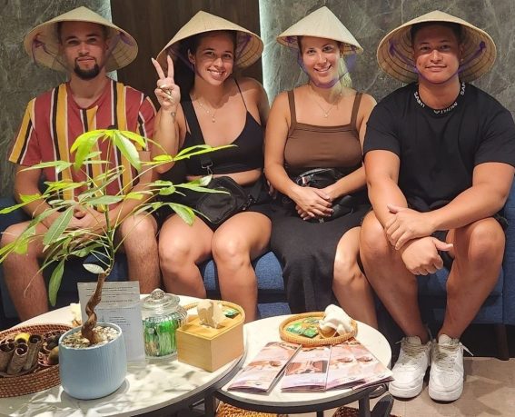 Two happy couples are smiling while wearing Vietnamese conical hats and sitting on a blue sofa in a spa.